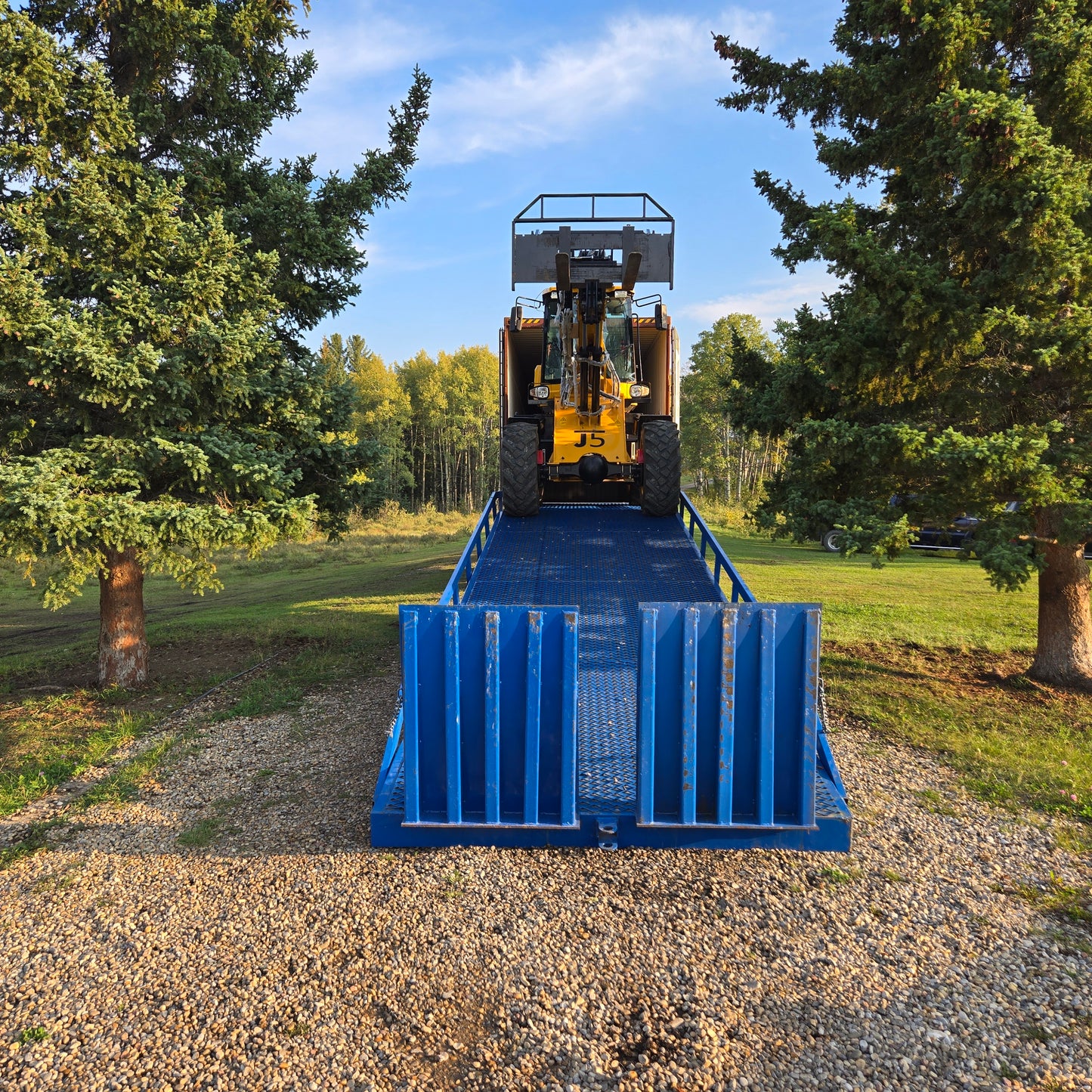 Yellow forklift front view driving up a blue twelve ton mobile loading ramp placed on gravel surface between green trees in Canada