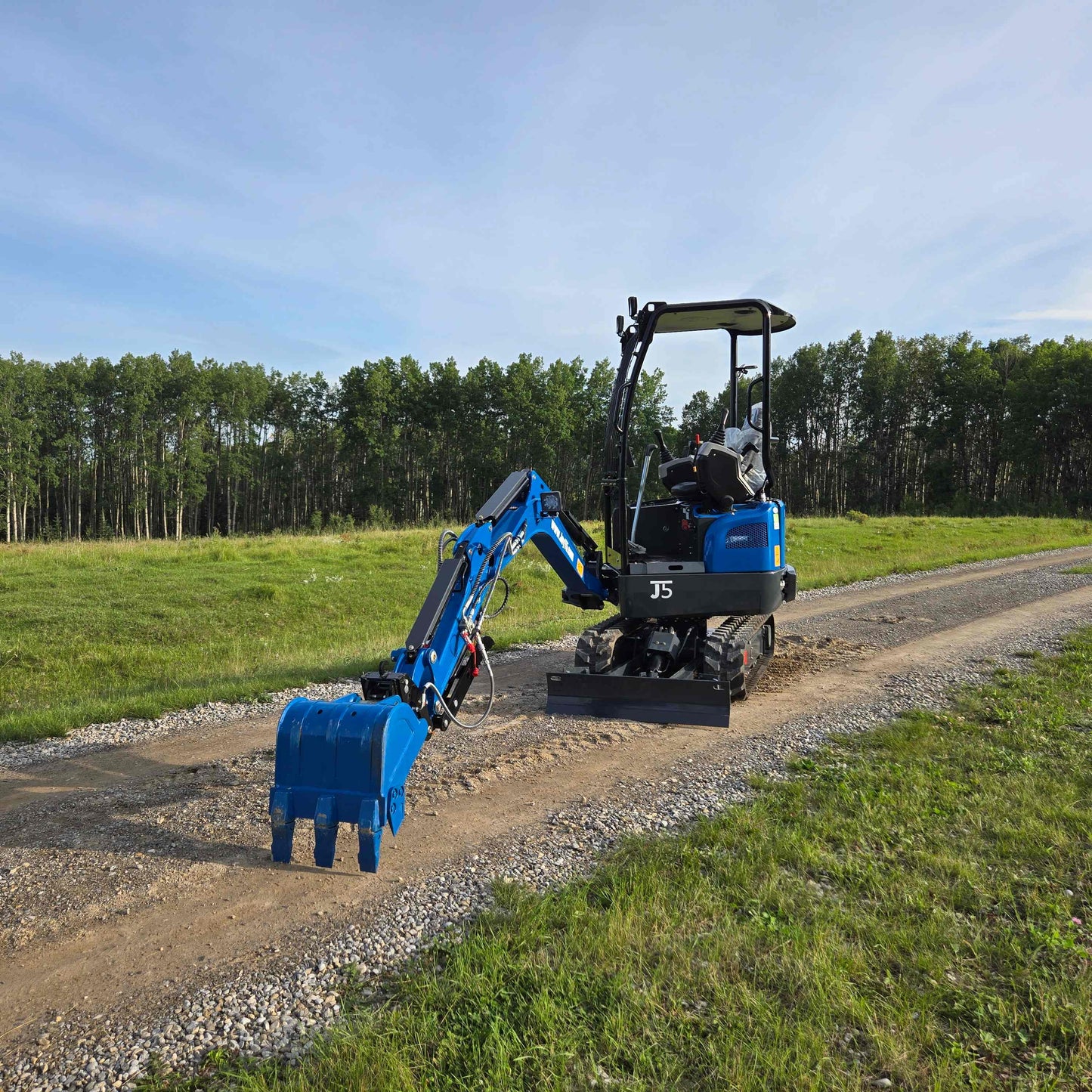 Back view of JoyT5 Rippa R18 Mini Excavator with digging bucket extended on a gravel road surrounded by green field and trees