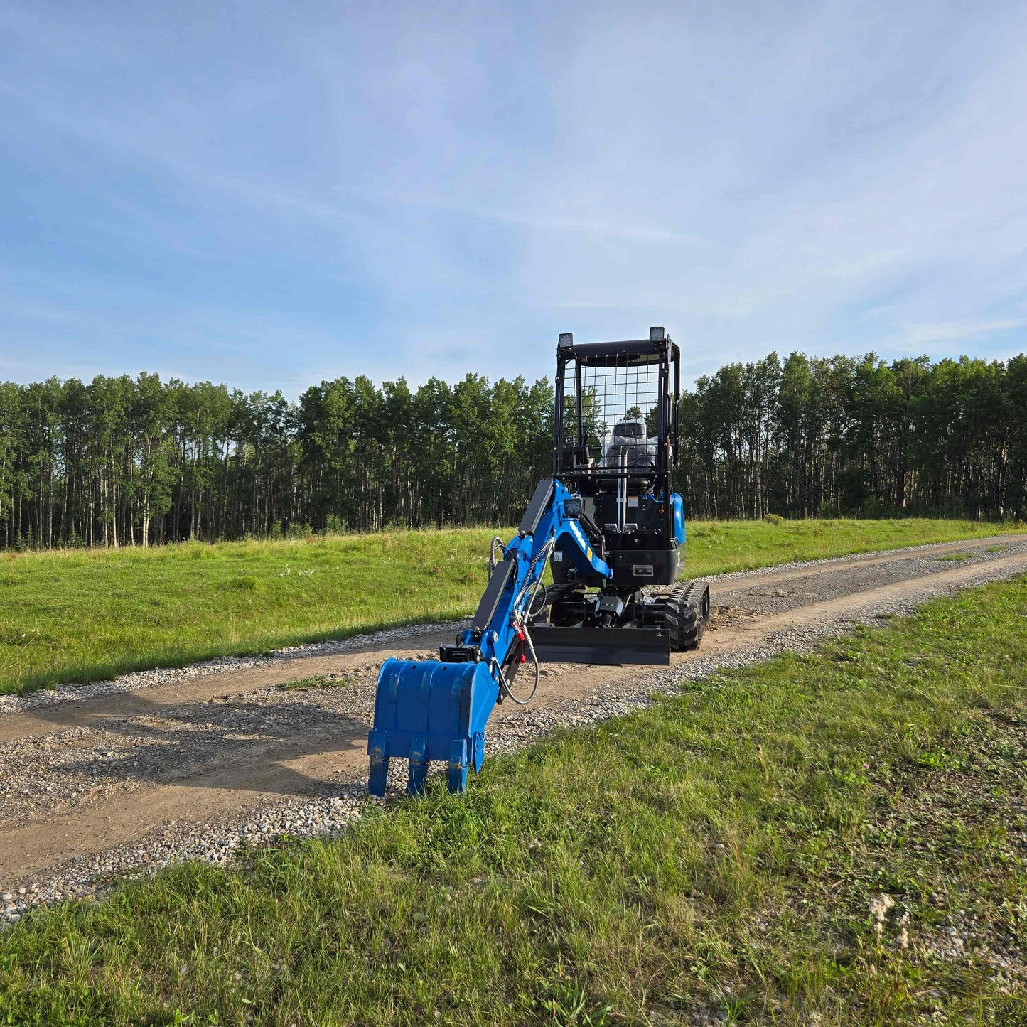 Back view of JoyT5 Rippa R18 Mini Excavator with blue digging bucket resting on gravel road beside green grass and trees.