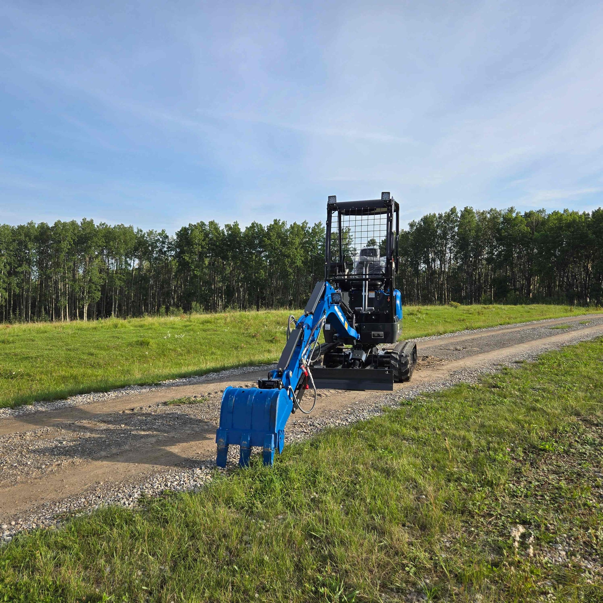 Back view of JoyT5 Rippa R18 Mini Excavator with blue digging bucket resting on gravel road beside green grass and trees.