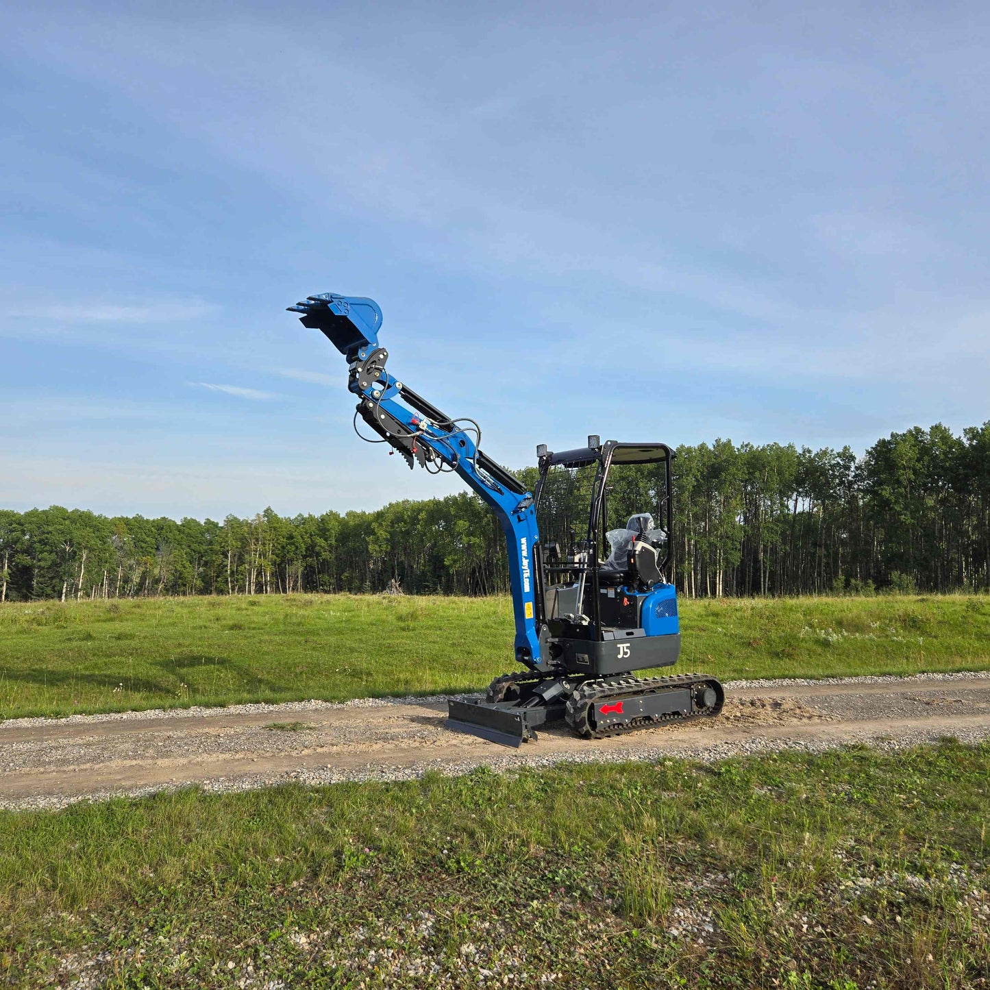 JoyT5 Rippa R18 Mini Excavator with bucket raised while working on a dirt road near a green field and trees