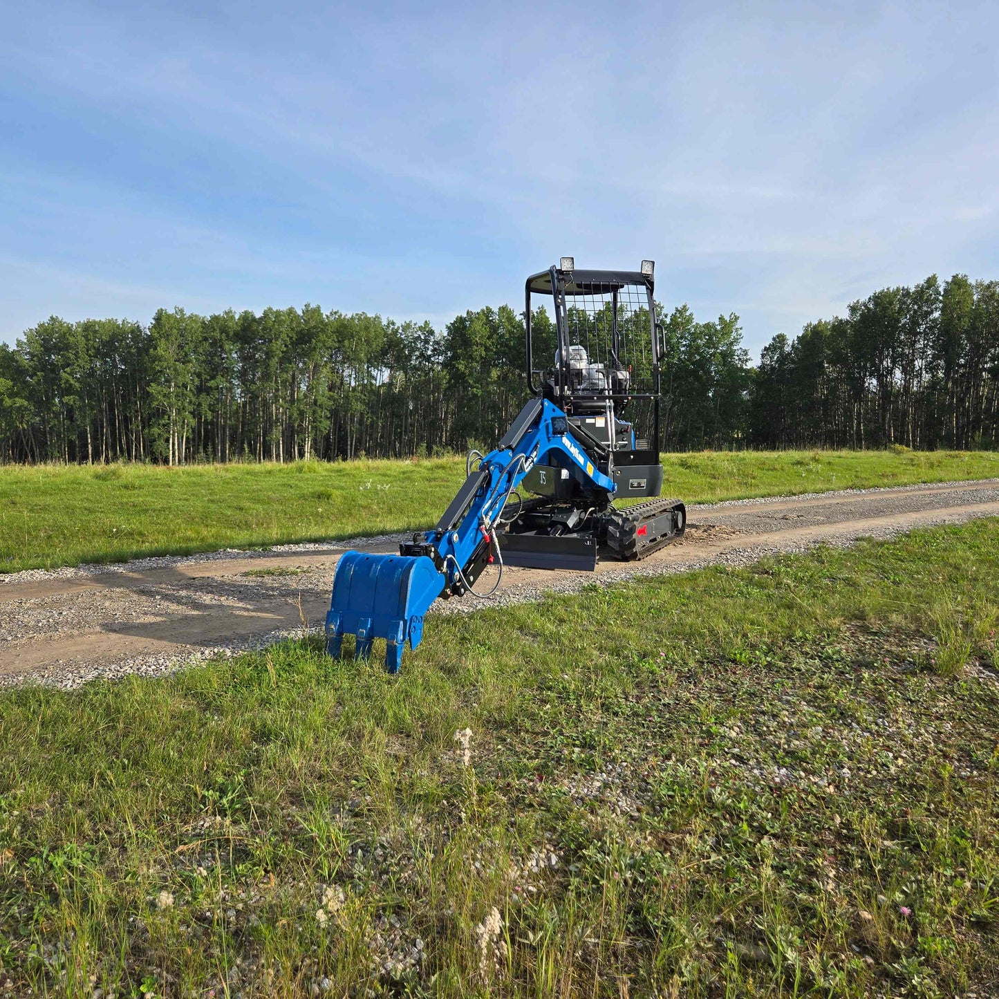 JoyT5 Rippa R18 Mini Excavator with extended digging arm and blue bucket on gravel road beside grassy field and forest background.