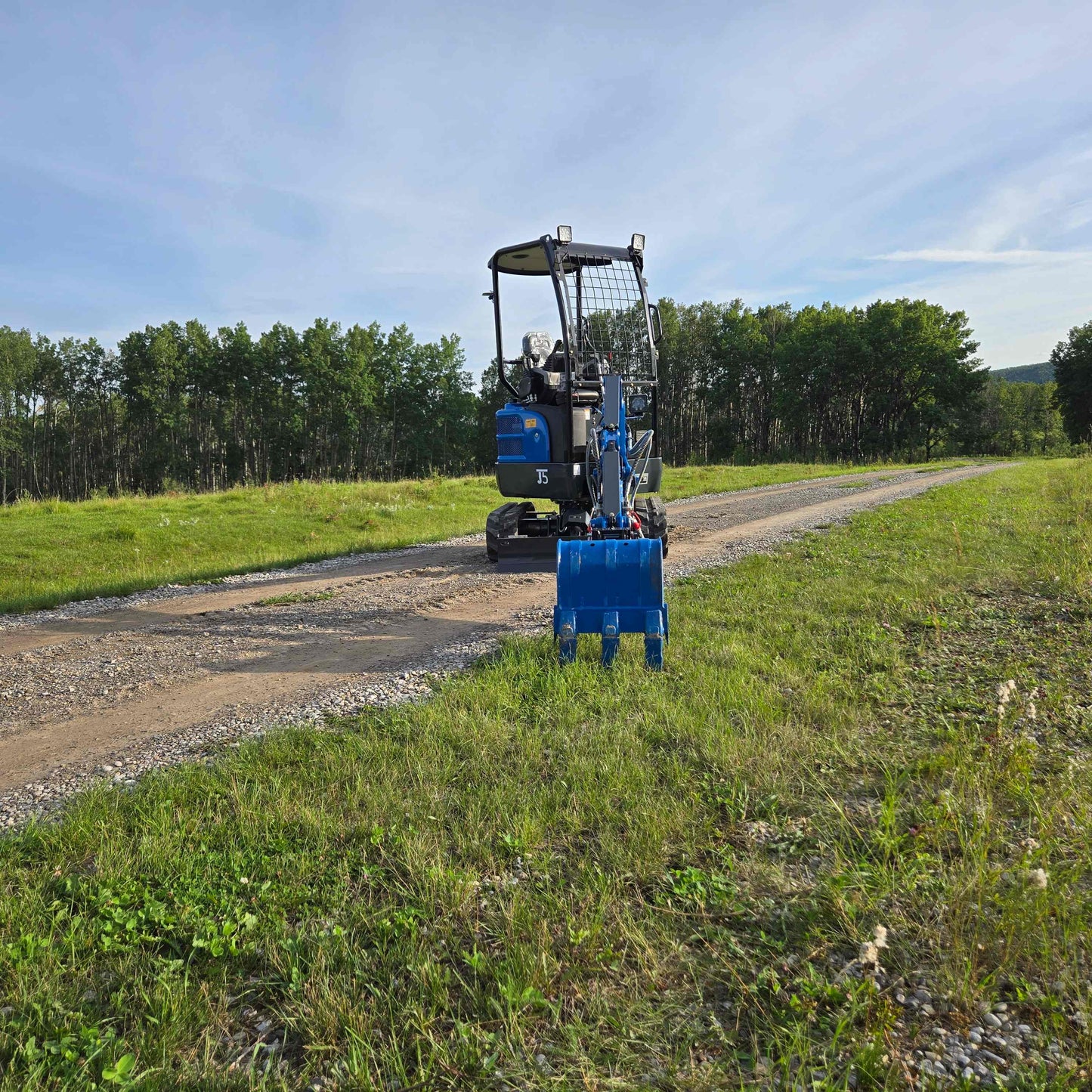 Front view of JoyT5 Rippa R18 Mini Excavator on a gravel road with its blue digging bucket extended over the grass, surrounded by trees and greenery