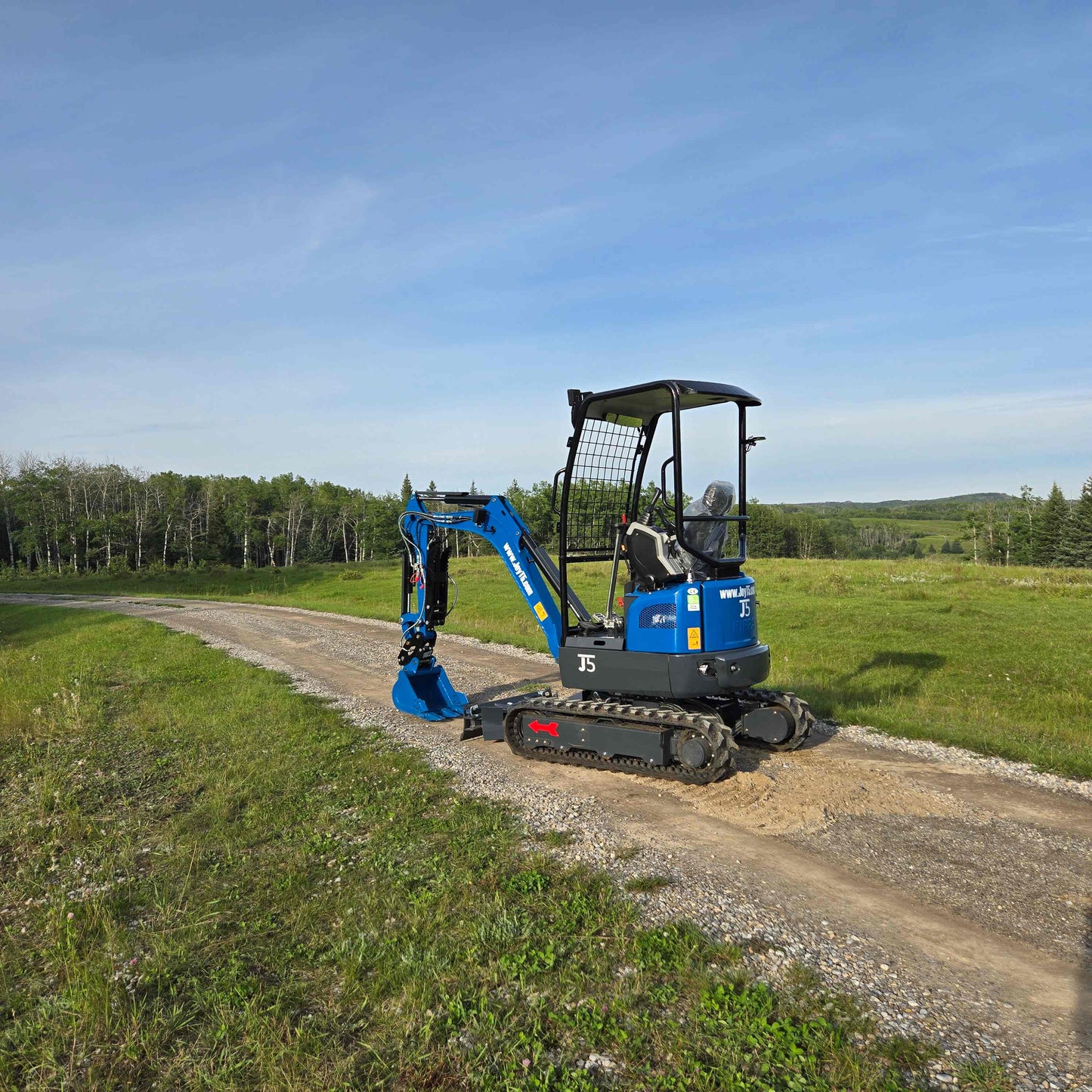JoyT5 Rippa R18 Mini Excavator working on a rural dirt road with bucket lowered beside green fields