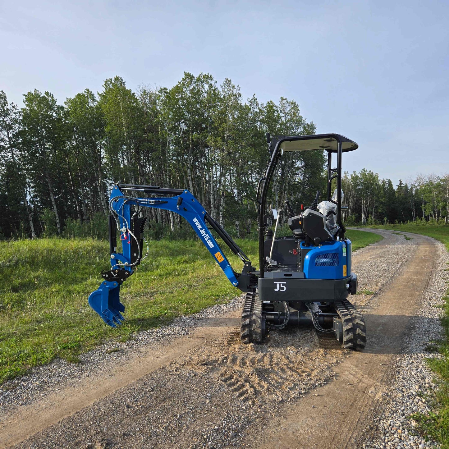 JoyT5 Rippa R18 Mini Excavator on a gravel road with its boom and bucket arm extended to the side, surrounded by green trees and grass
