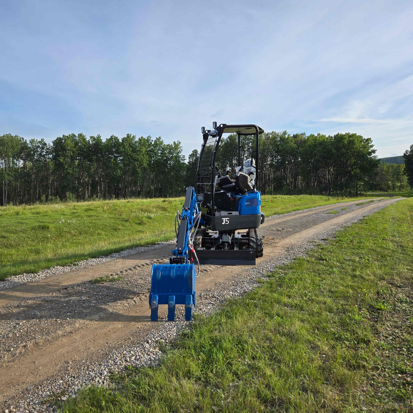 Rear view of JoyT5 Rippa R18 Mini Excavator on a gravel road with digging bucket extended forward and green landscape in the background
