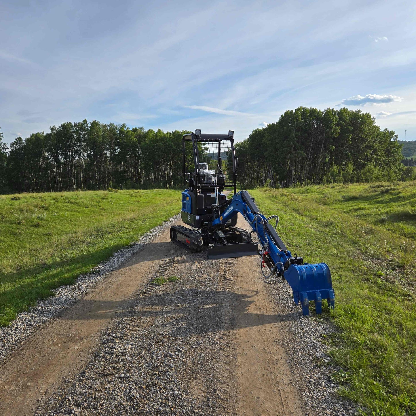 Rear view of JoyT5 Rippa R18 Mini Excavator with extended blue digging arm and bucket placed on gravel road in a green field with trees