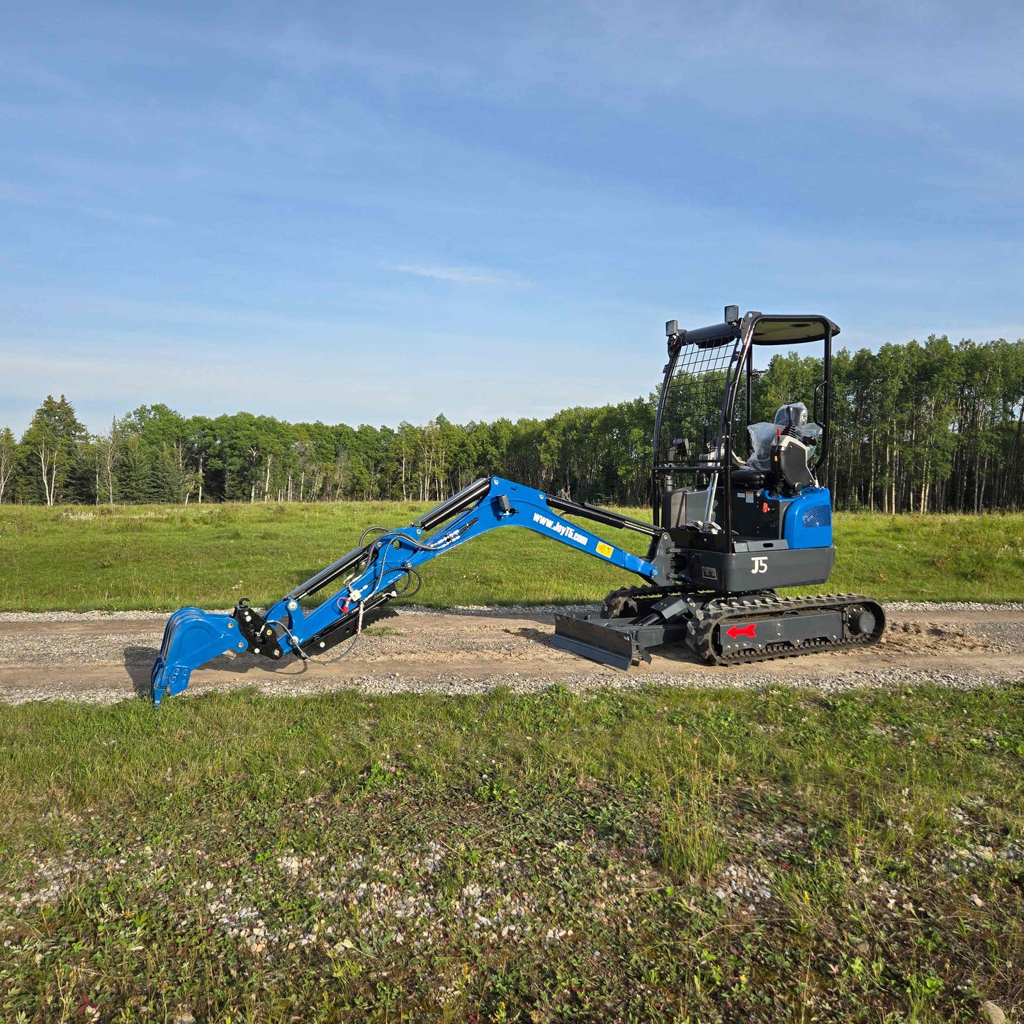 Side view of JoyT5 Rippa R18 Mini Excavator with extended digging arm and bucket resting on gravel road beside a grassy field and trees