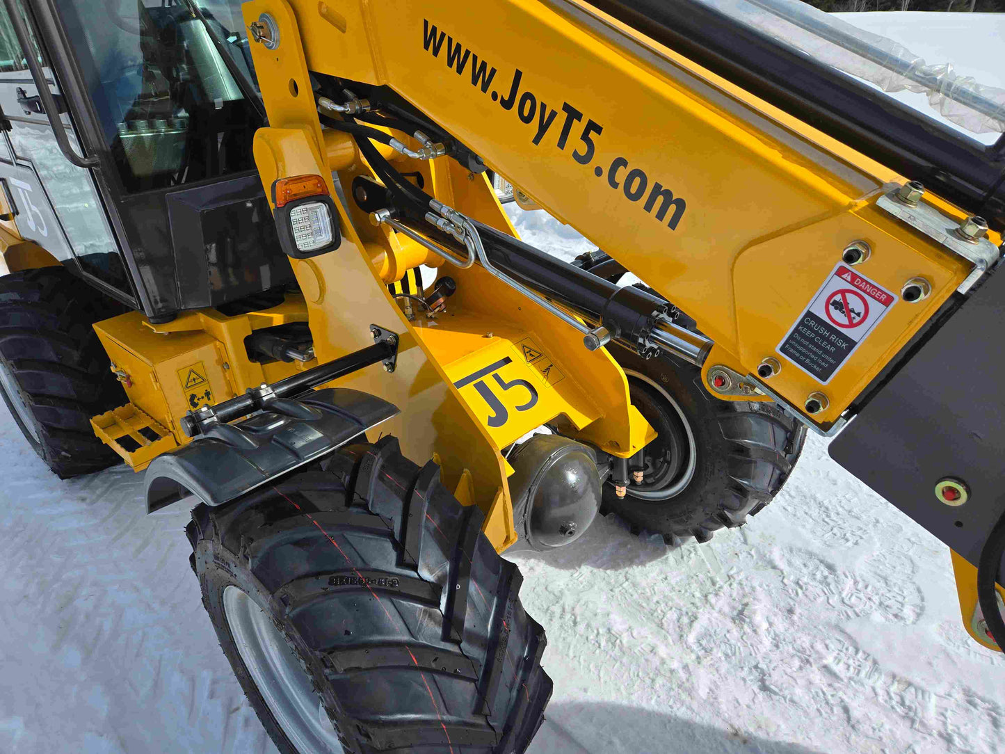 JT5WL1000 mini wheel loader arm showing hydraulic system, safety warning label, and large durable tires on snow.