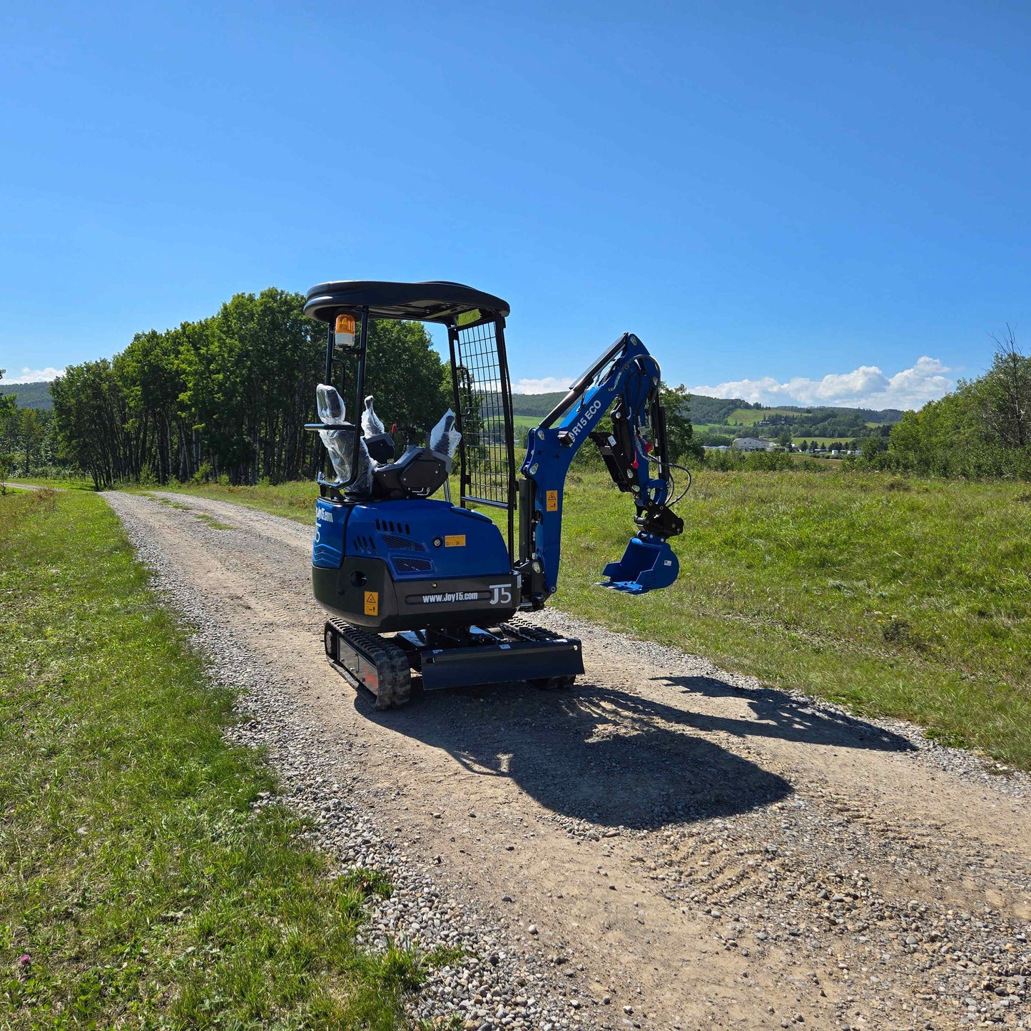 Rippa R15 Mini Excavator For Sale – compact blue excavator positioned on gravel road with green field, trees, and cloudy sky in the background.