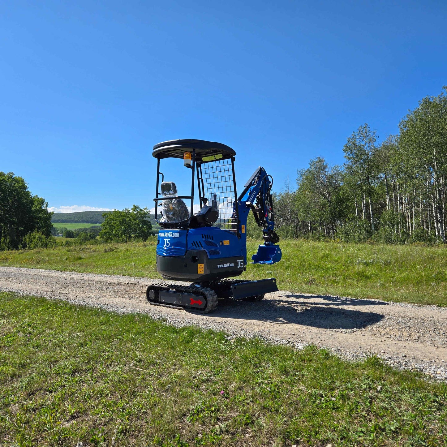 Rippa R15 Mini Excavator For Sale – side view of compact excavator on gravel road with grassy field and trees in the background.