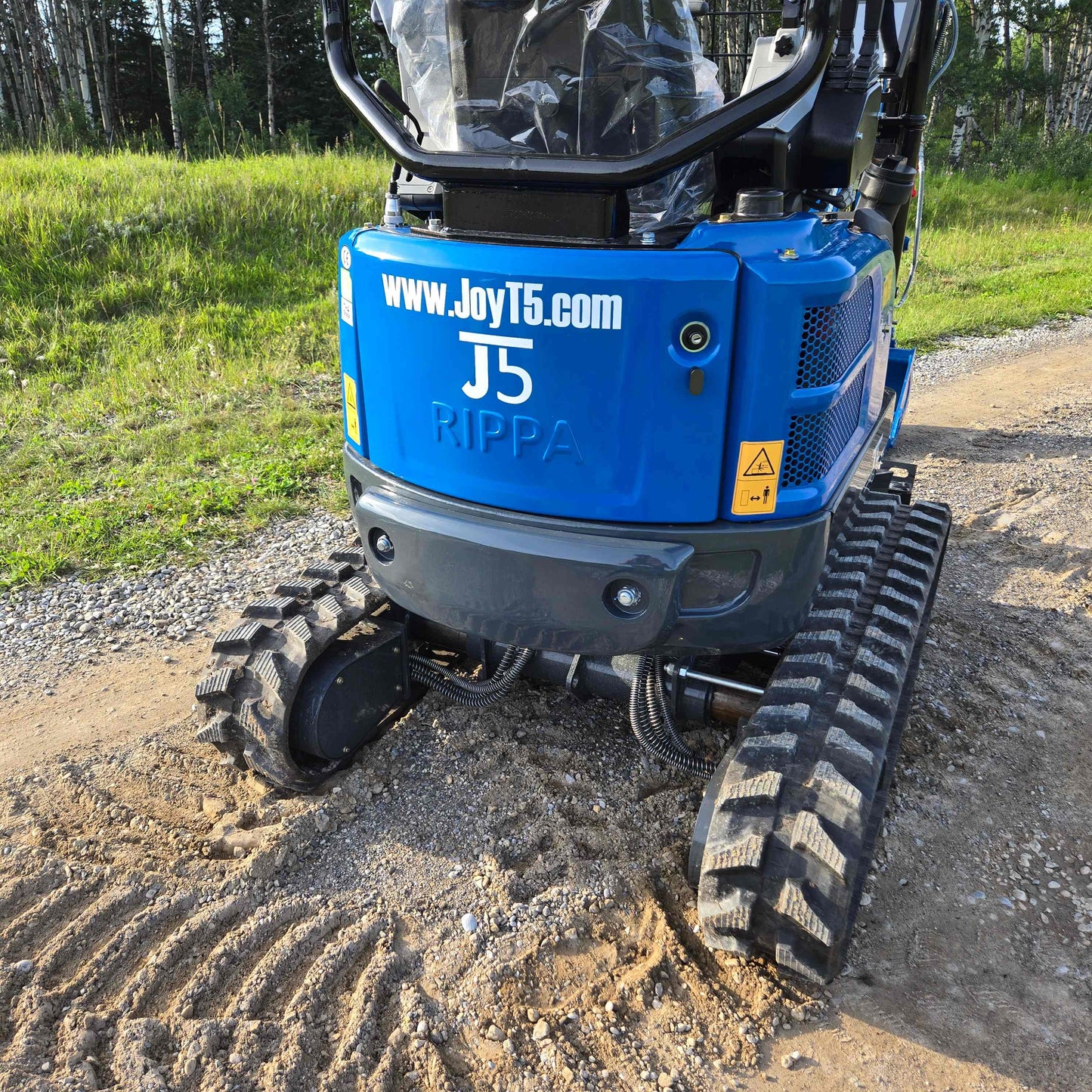 Rear view of JoyT5 Rippa R18 Mini Excavator for sale with branding and rubber tracks visible, positioned on a gravel and dirt road surrounded by greenery.