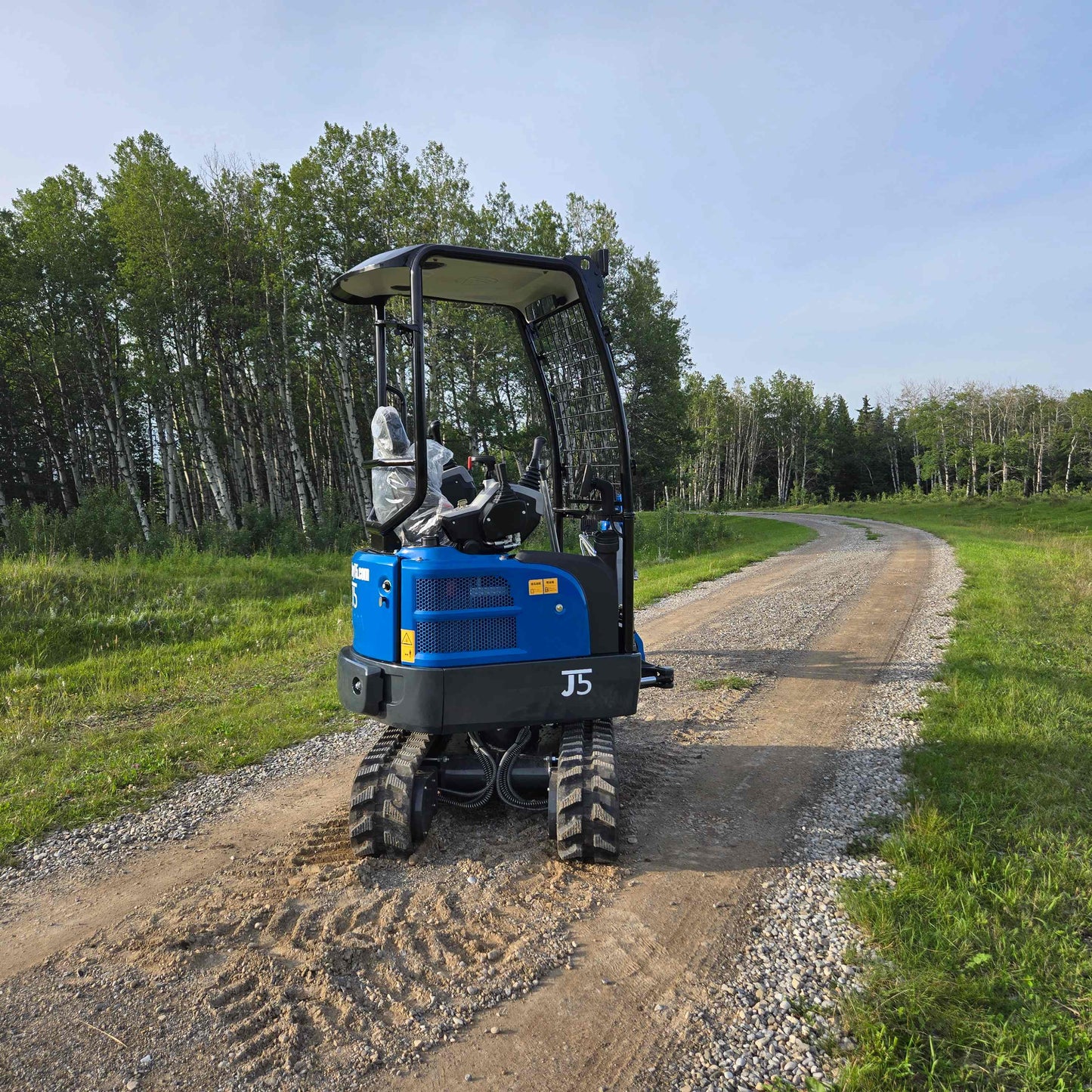 Rear view of JoyT5 Rippa R18 Mini Excavator parked on a gravel road with grass field and trees in the background