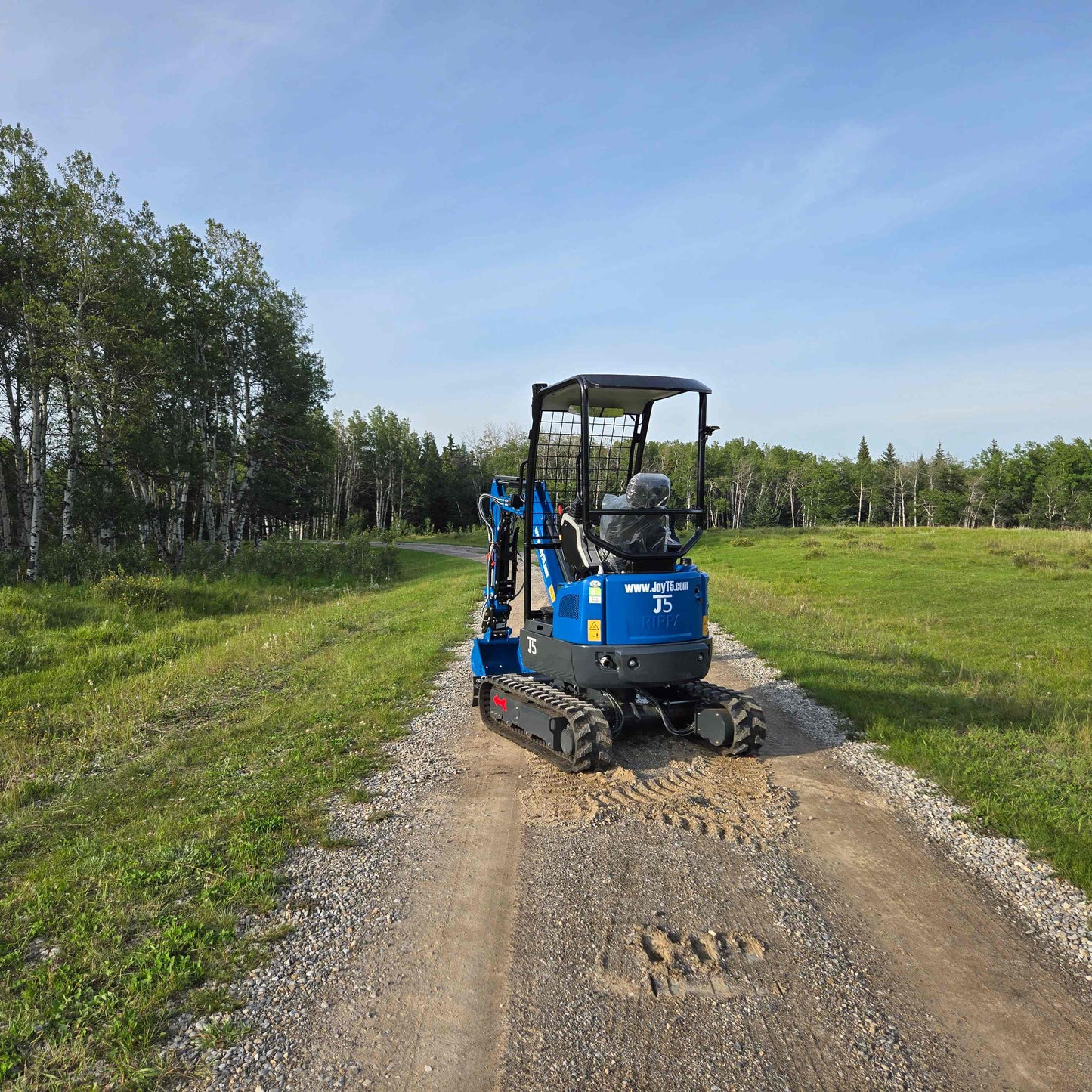 Rear view of JoyT5 Rippa R18 Mini Excavator on gravel road with green grass and forest background