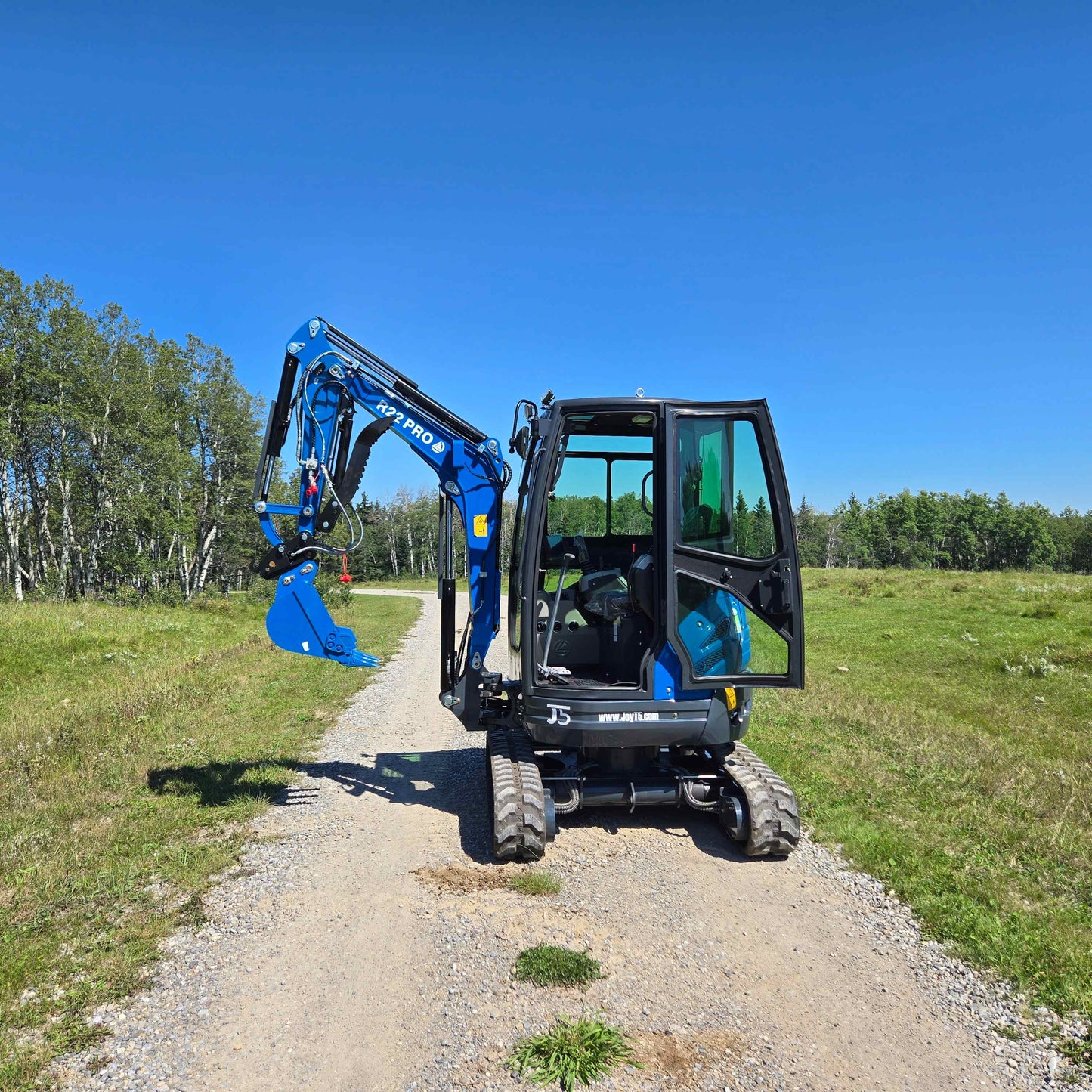 Front view of the Rippa R22 Mini Excavator for sale with cabin door open and digging arm extended, parked on a gravel path surrounded by grass and trees.