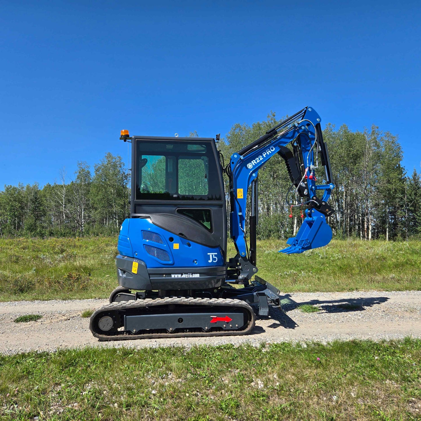 Full side view of Rippa R22 Mini Excavator for sale on gravel road with bucket attachment raised, in front of trees.