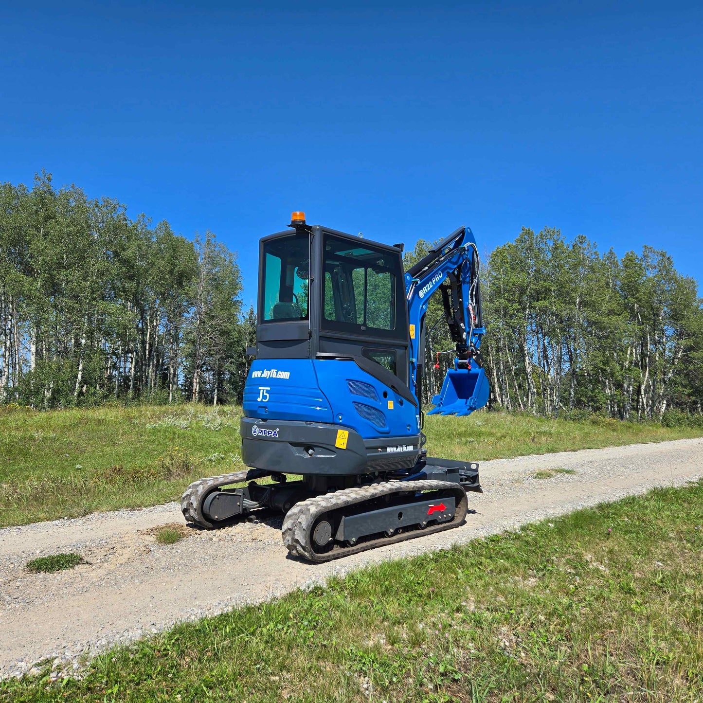 Rear side view of Rippa R22 Mini Excavator for sale on gravel road with bucket lowered, trees in the background.
