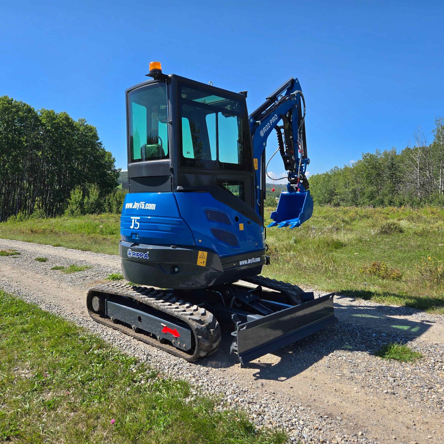 Rear side view of the Rippa R22 Mini Excavator for sale with front blade and digging arm extended, parked on a gravel path with trees in the background.