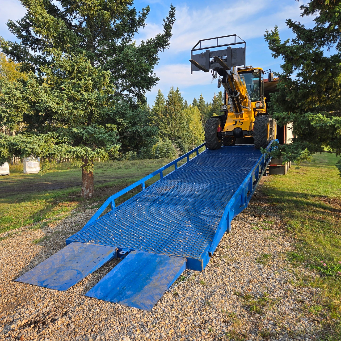 Yellow forklift driving down blue steel twelve ton mobile loading ramp on gravel road surrounded by green trees used for truck loading in Canada

