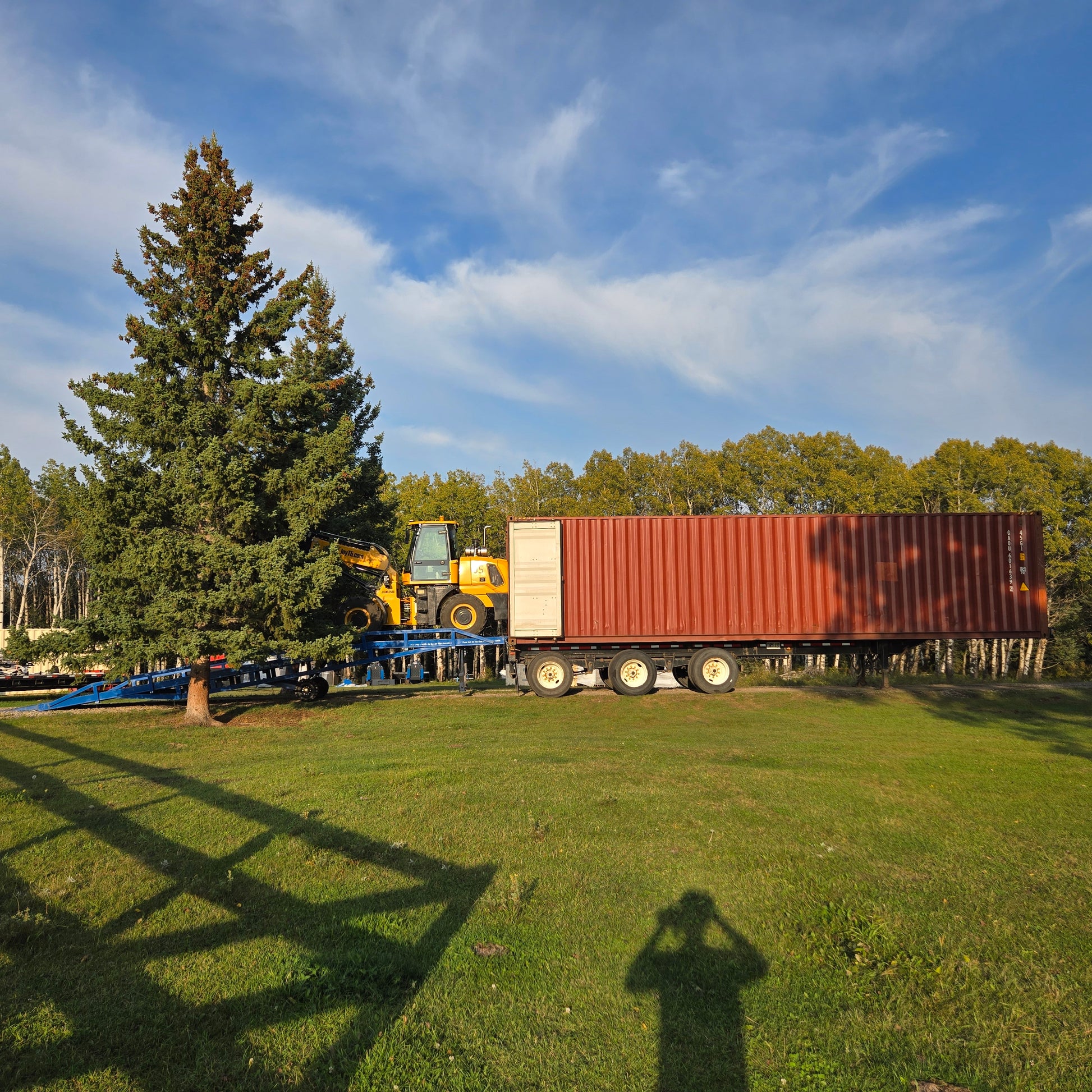 Yellow forklift loading a large container truck using a blue twelve ton mobile loading ramp on an open grass field in Canada under clear blue sky

