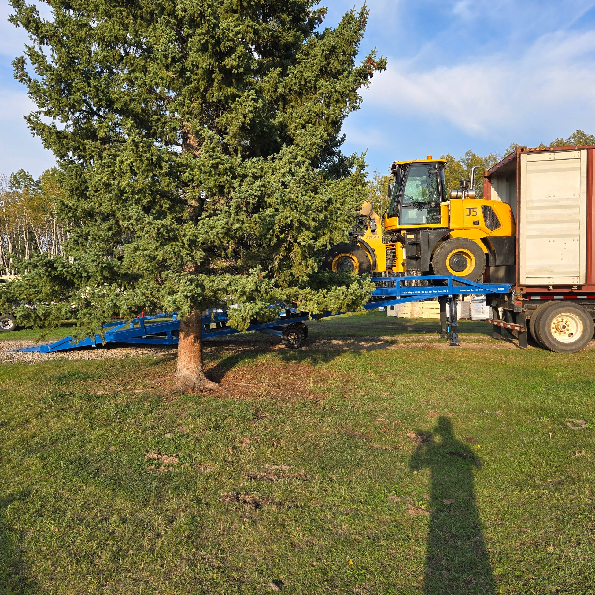 Yellow forklift loading a shipping container truck using a blue twelve ton mobile loading ramp in a grassy yard area surrounded by trees in Canada

