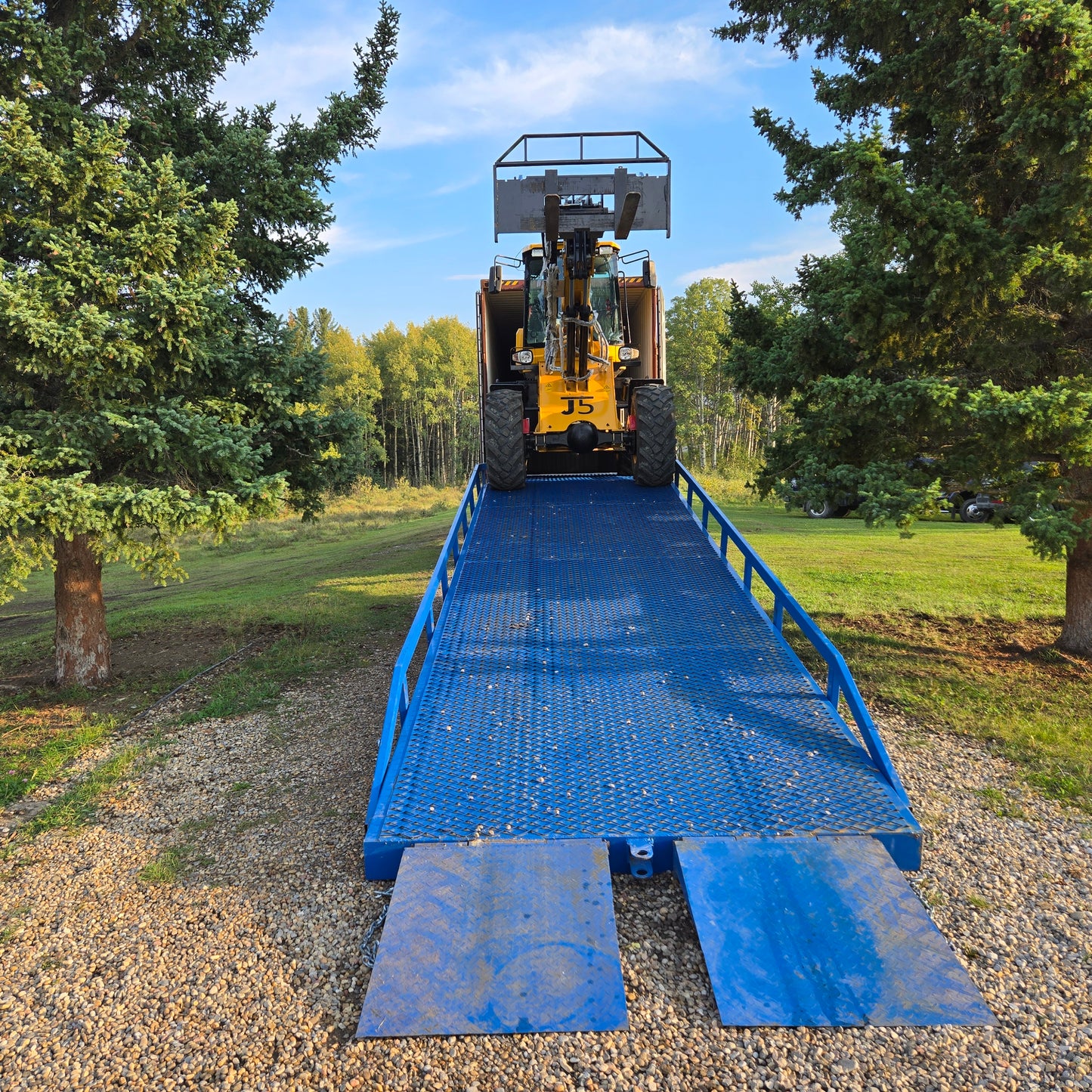 Yellow forklift driving up twelve ton blue steel mobile loading ramp surrounded by green trees used for truck and container loading in Canada


