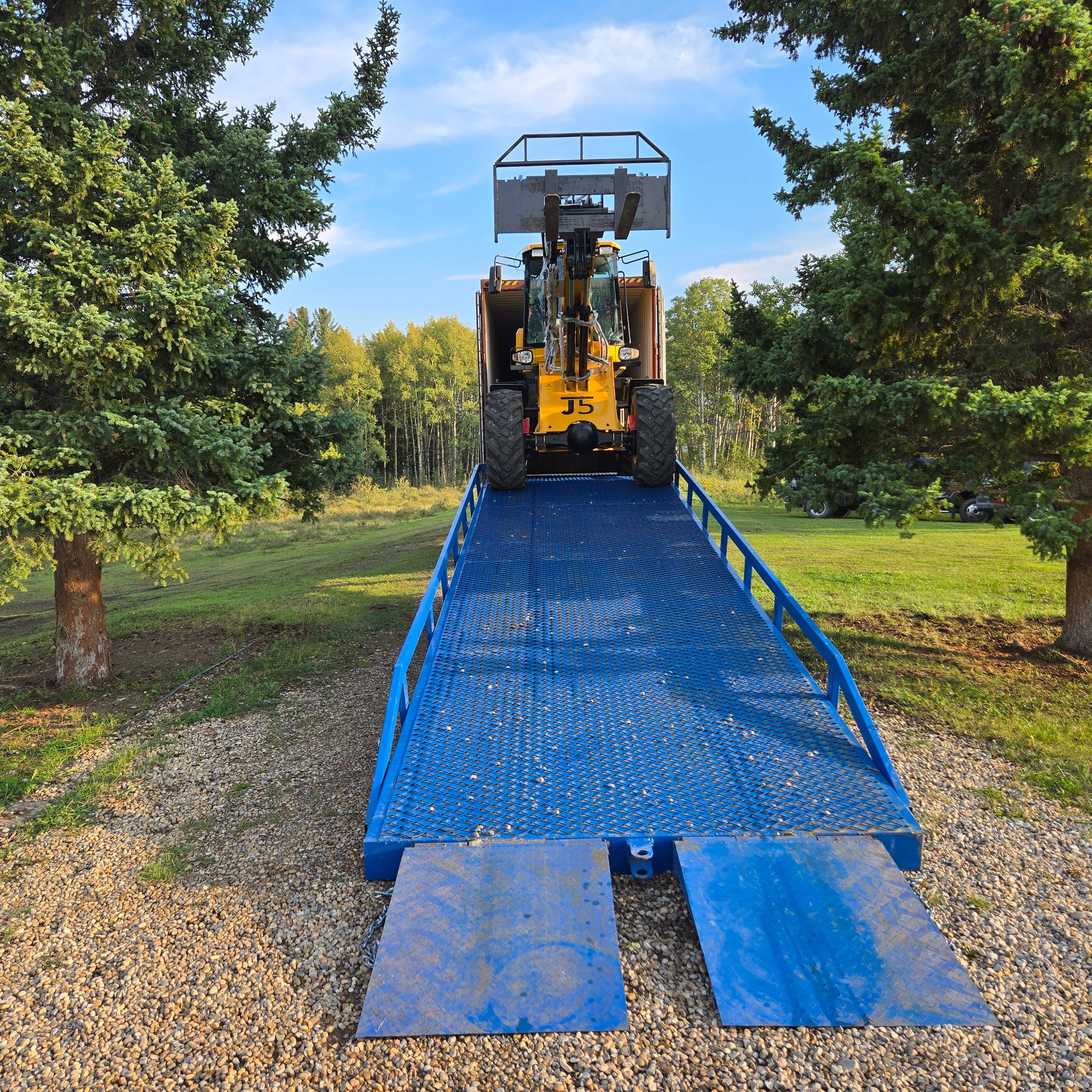 Yellow forklift driving up twelve ton blue steel mobile loading ramp surrounded by green trees used for truck and container loading in Canada

