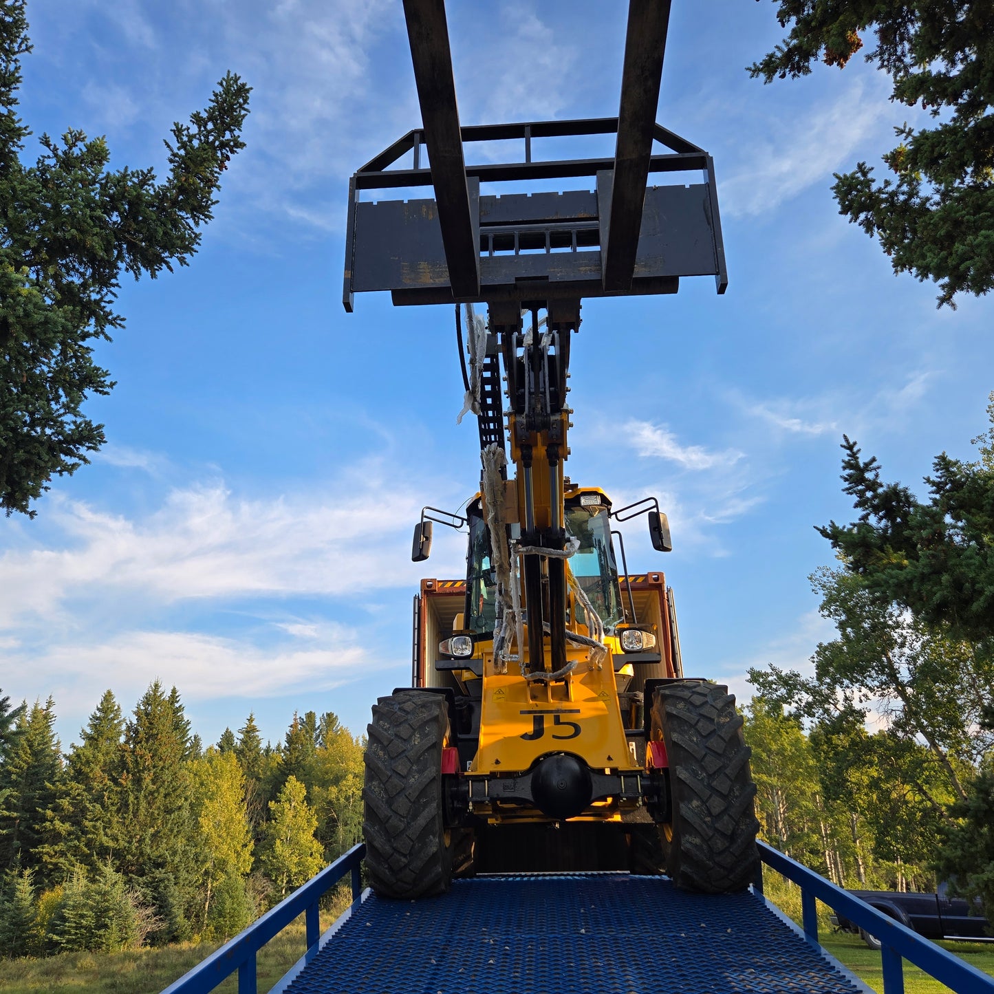 Yellow forklift front view driving up blue twelve ton mobile loading ramp with raised bucket against clear sky and green trees in Canada

