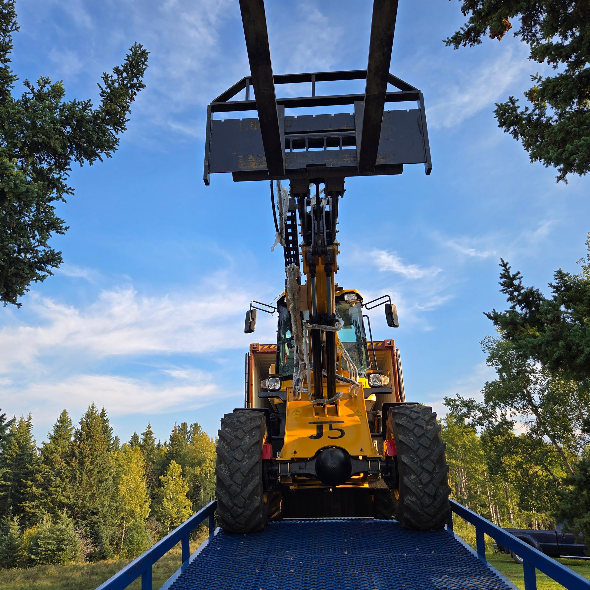 Yellow forklift front view driving up blue twelve ton mobile loading ramp with raised bucket against clear sky and green trees in Canada

