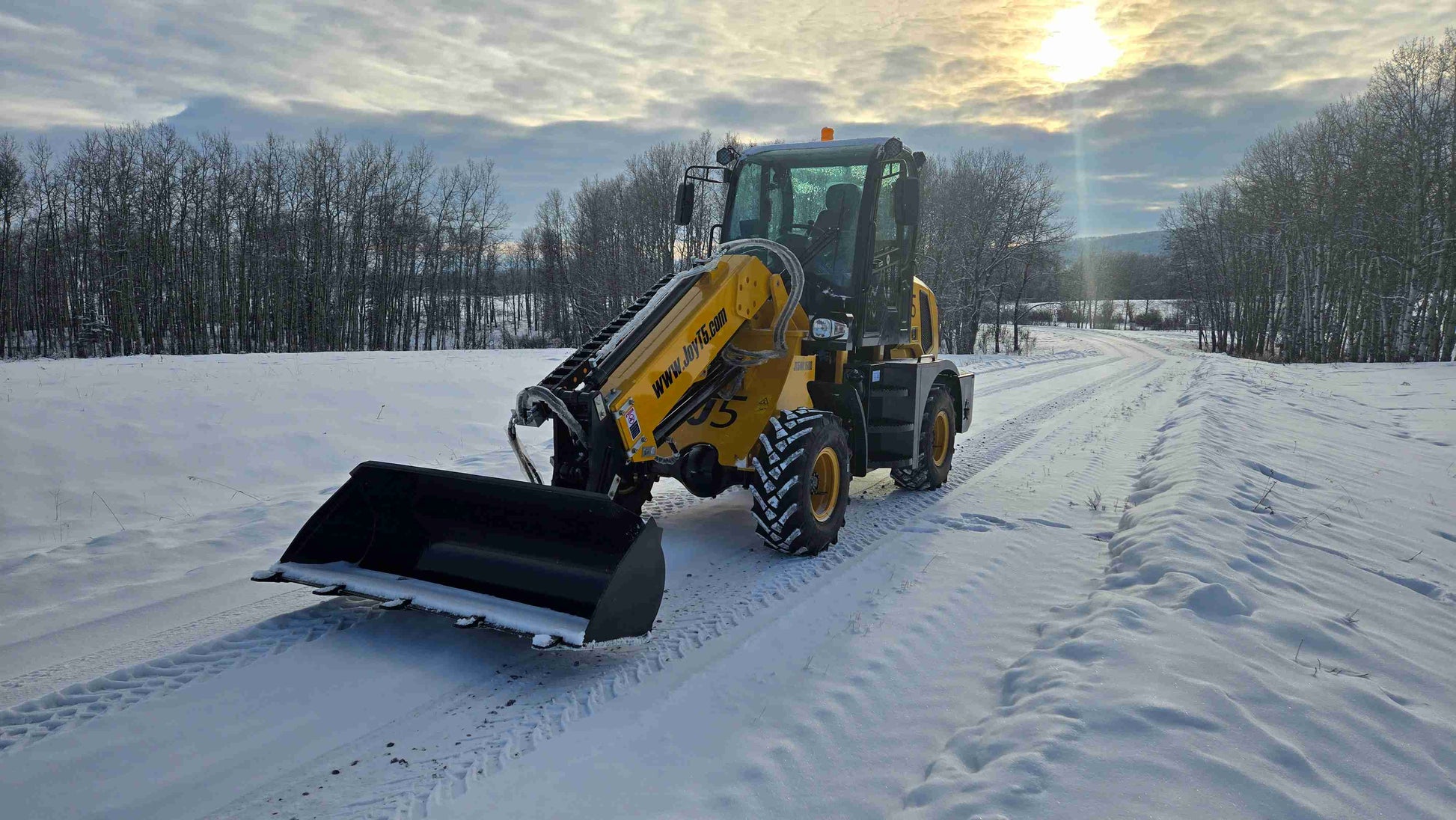 JT5WL1500 compact wheel loader front view with bucket resting on snowy road under sunset sky.