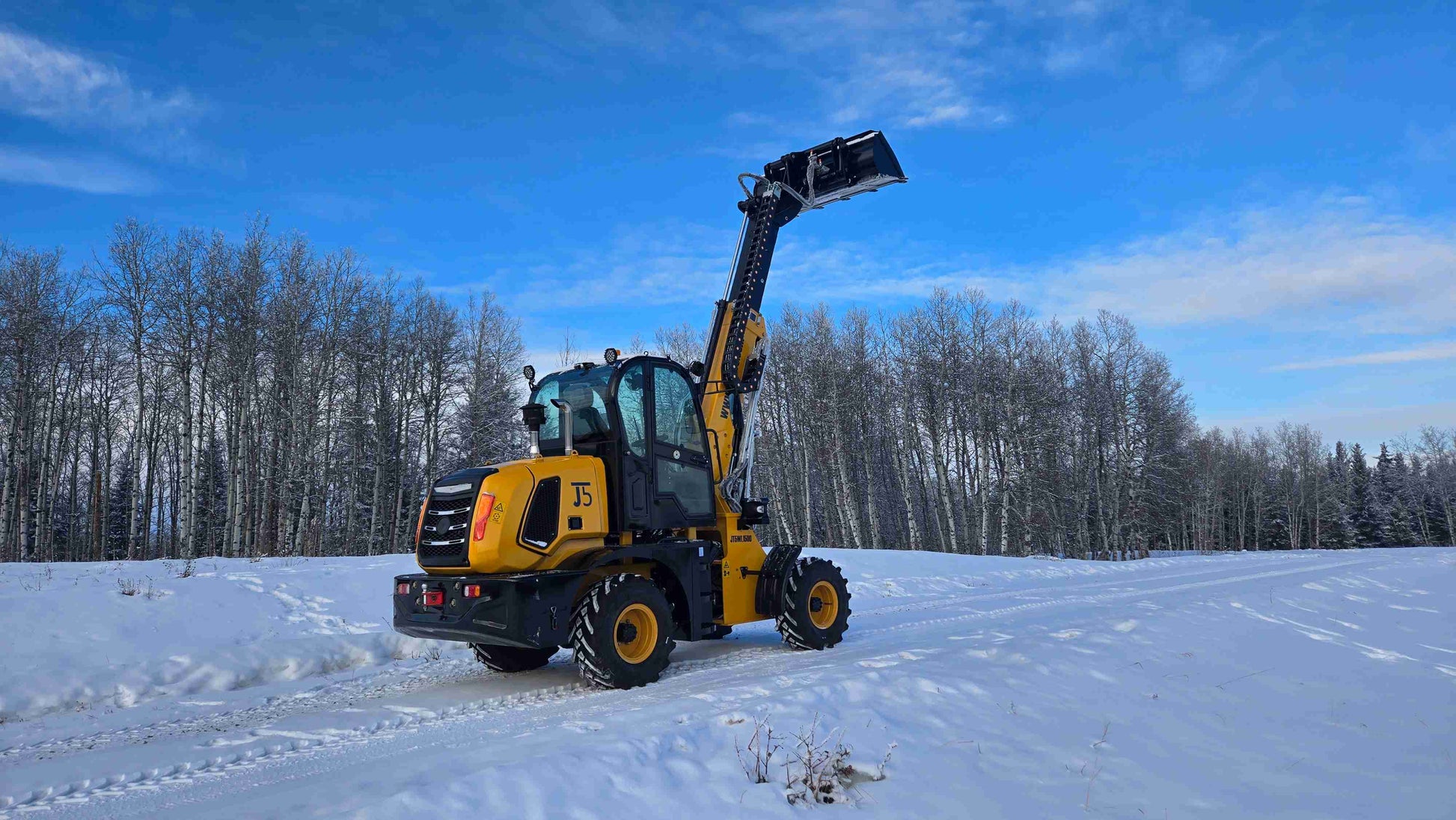 JT5WL1500 wheel loader rear angle with extended boom and raised bucket on snowy field with forest background.