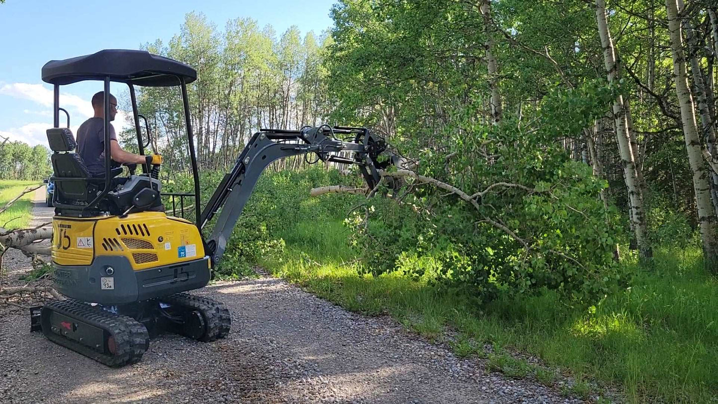 J5 mini excavator with tree grab attachment lifting and moving a fallen tree branch in a forest area.
