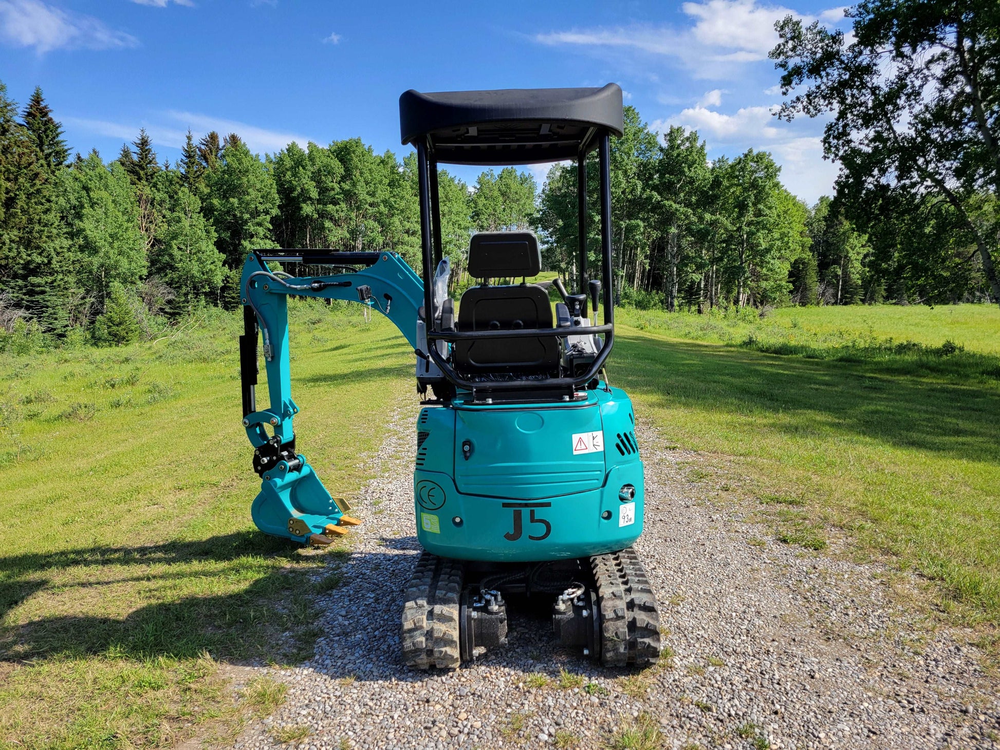 Rear view of JoyT5 mini excavator showing operator seat, protective canopy, rubber tracks, and digging arm in a field setting.
