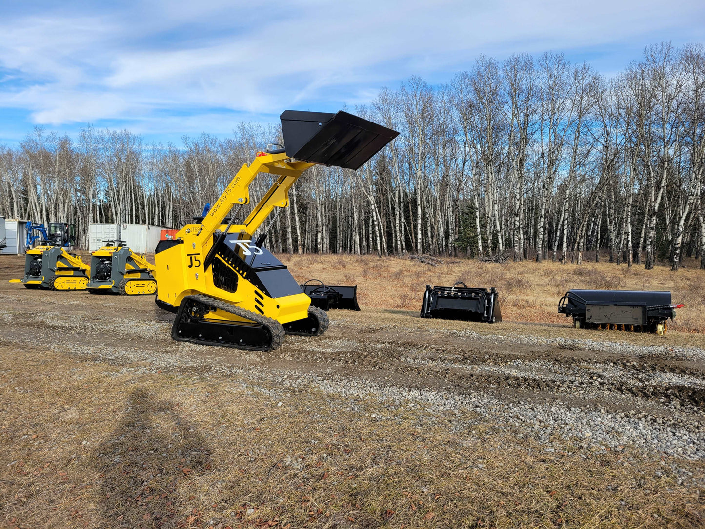 A JoyT5 mini skid steer loader in Canada lifting a bucket attachment with multiple JoyT5 machines and attachments displayed in an outdoor work area
