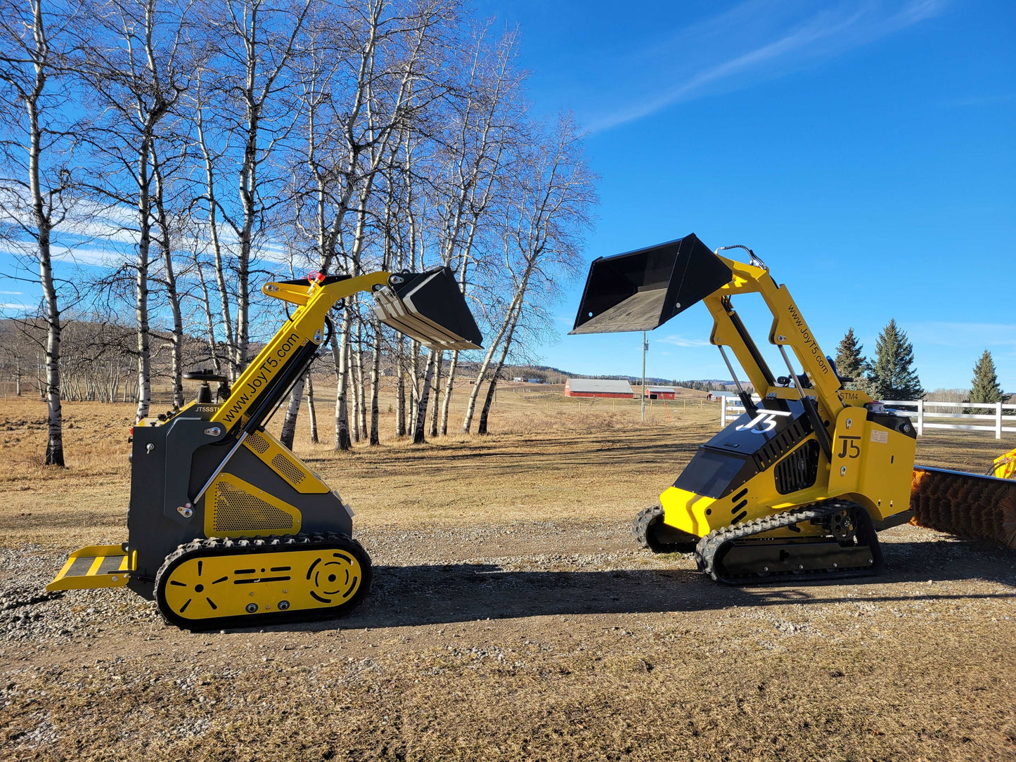 Two JoyT5 mini skid steer loaders side by side, one tracked and one wheeled, both with raised buckets outdoors.