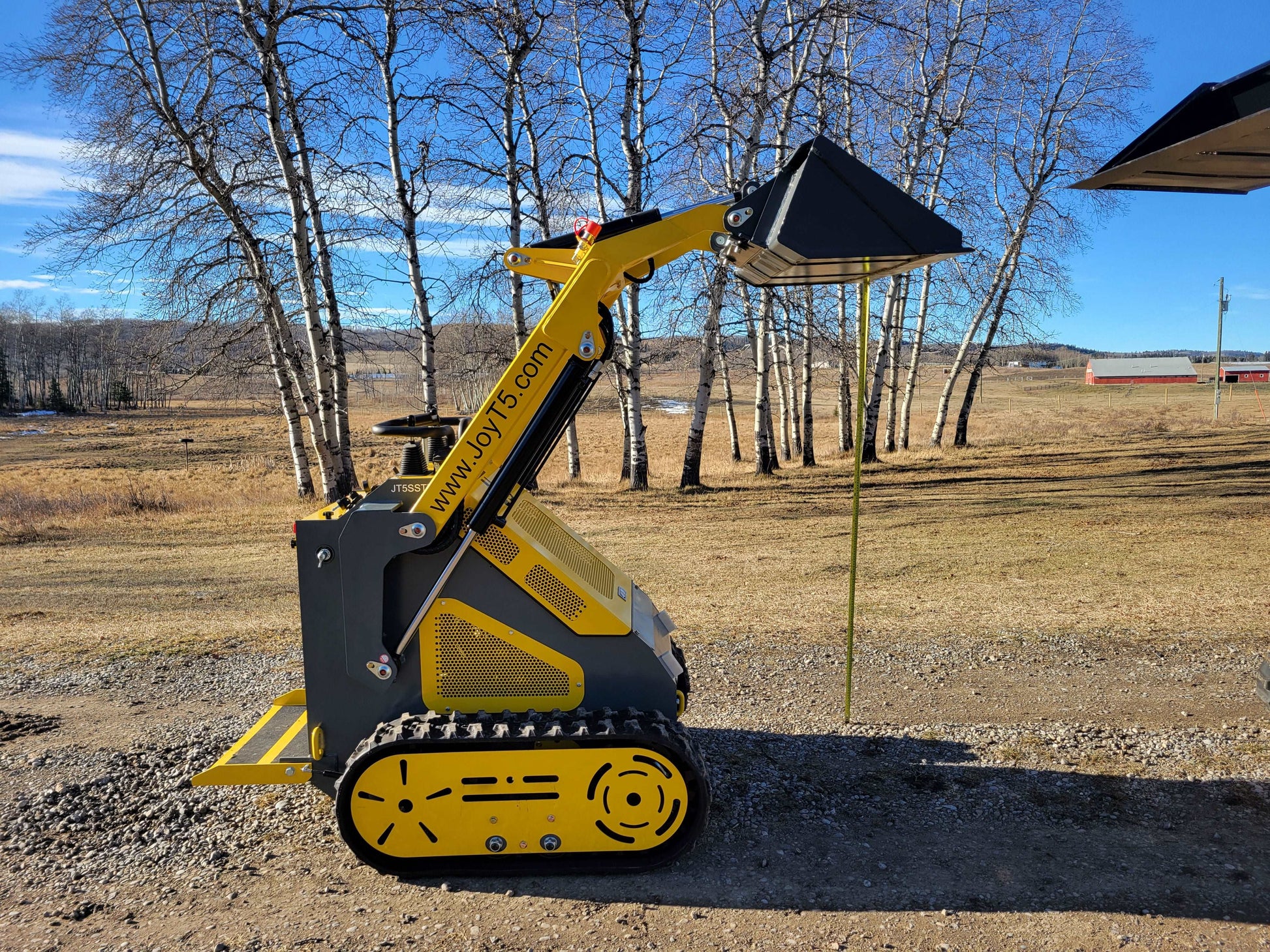 JoyT5 mini skid steer loader demonstrating maximum bucket lift height outdoors on gravel and grass terrain.