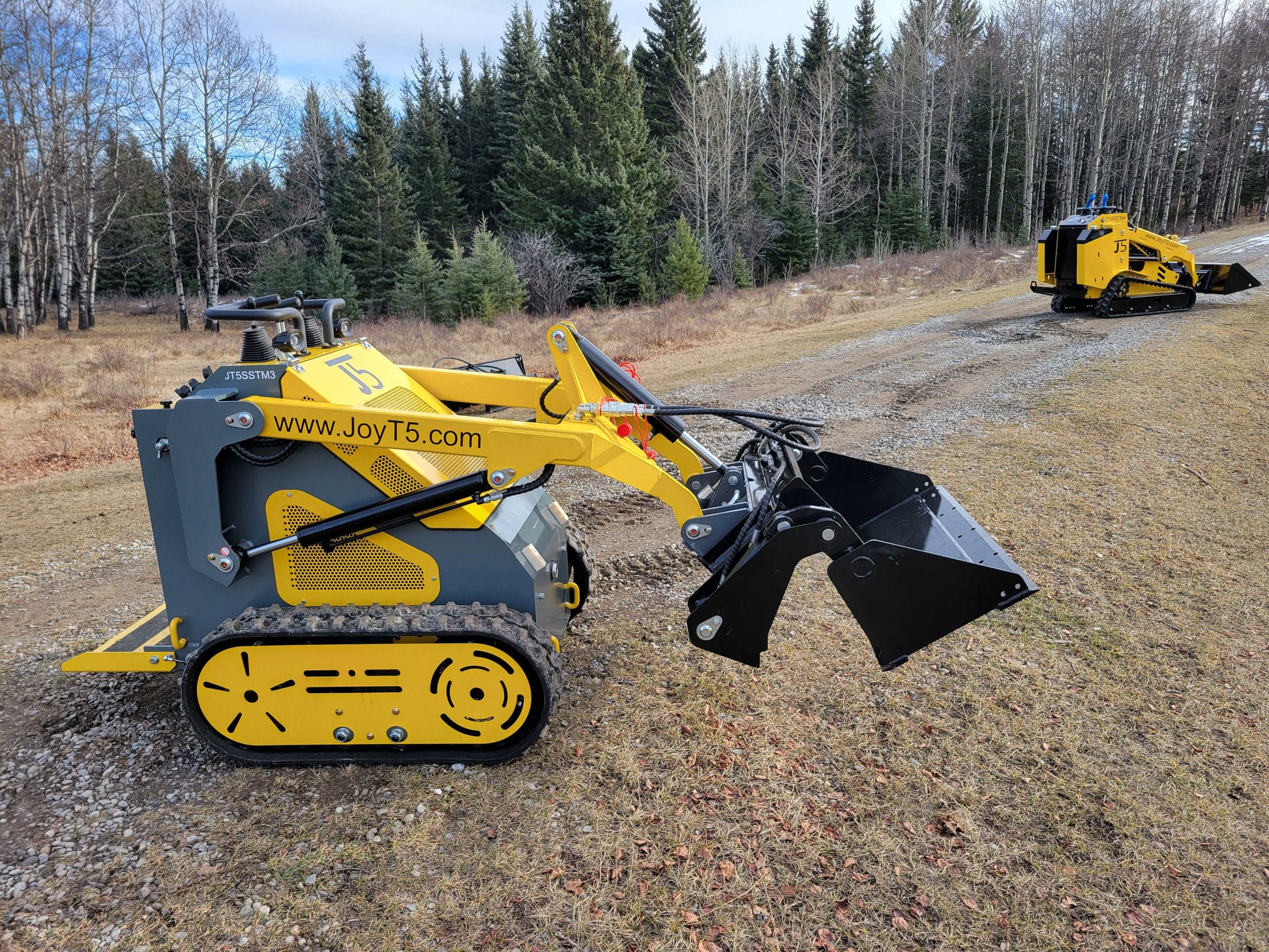Compact JoyT5 mini skid steer with bucket attachment on gravel road, with another skid steer in the background.