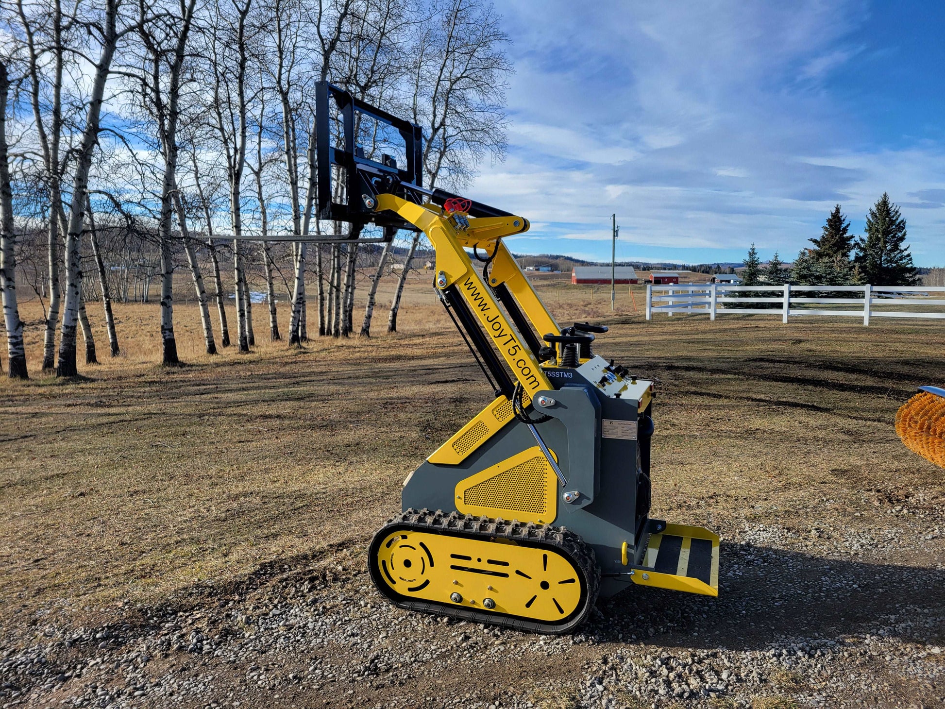 JoyT5 mini tracked skid steer with raised fork attachment shown from the side in an outdoor Canada work area