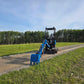 Back view of JoyT5 Rippa R18 Mini Excavator with blue digging bucket resting on gravel road beside green grass and trees.