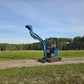 JoyT5 Rippa R18 Mini Excavator with bucket raised while working on a dirt road near a green field and trees
