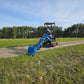 JoyT5 Rippa R18 Mini Excavator with extended digging arm and blue bucket on gravel road beside grassy field and forest background.