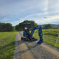 JoyT5 Rippa R18 Mini Excavator front view parked on dirt road with green field and hills in background