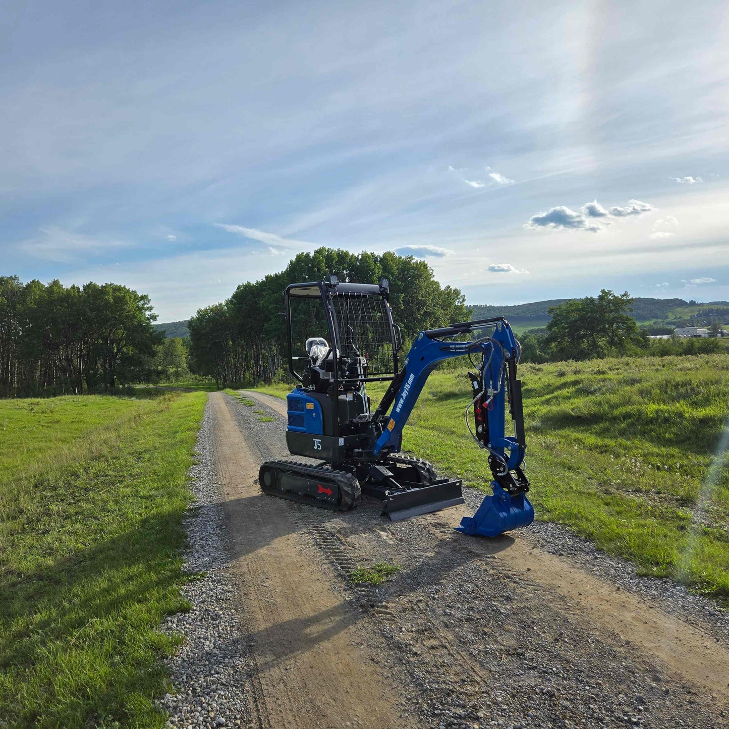 JoyT5 Rippa R18 Mini Excavator front view parked on dirt road with green field and hills in background