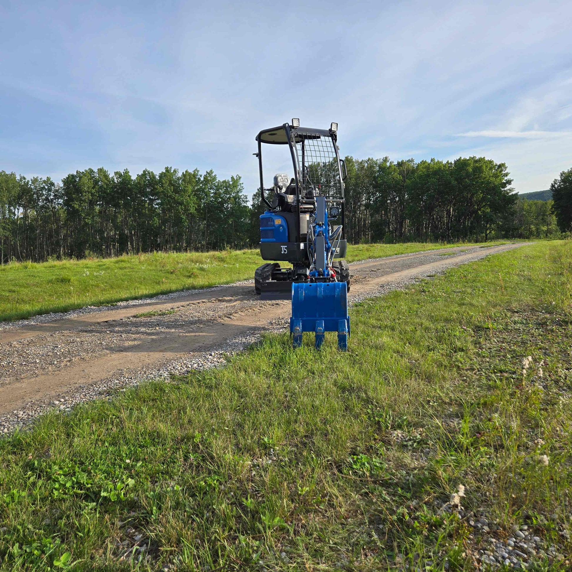Front view of JoyT5 Rippa R18 Mini Excavator on a gravel road with its blue digging bucket extended over the grass, surrounded by trees and greenery