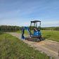 JoyT5 Rippa R18 Mini Excavator working on a rural dirt road with bucket lowered beside green fields