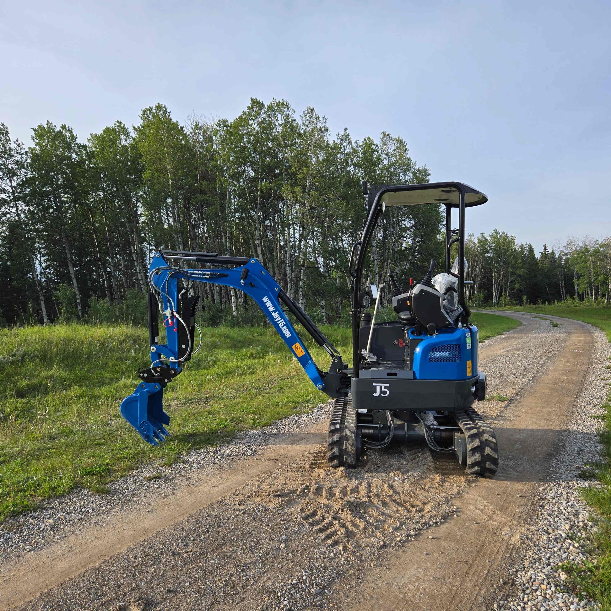 JoyT5 Rippa R18 Mini Excavator on a gravel road with its boom and bucket arm extended to the side, surrounded by green trees and grass
