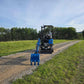 Rear view of JoyT5 Rippa R18 Mini Excavator on a gravel road with digging bucket extended forward and green landscape in the background