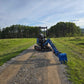 Rear view of JoyT5 Rippa R18 Mini Excavator with extended blue digging arm and bucket placed on gravel road in a green field with trees