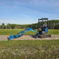 Side view of JoyT5 Rippa R18 Mini Excavator with extended digging arm and bucket resting on gravel road beside a grassy field and trees