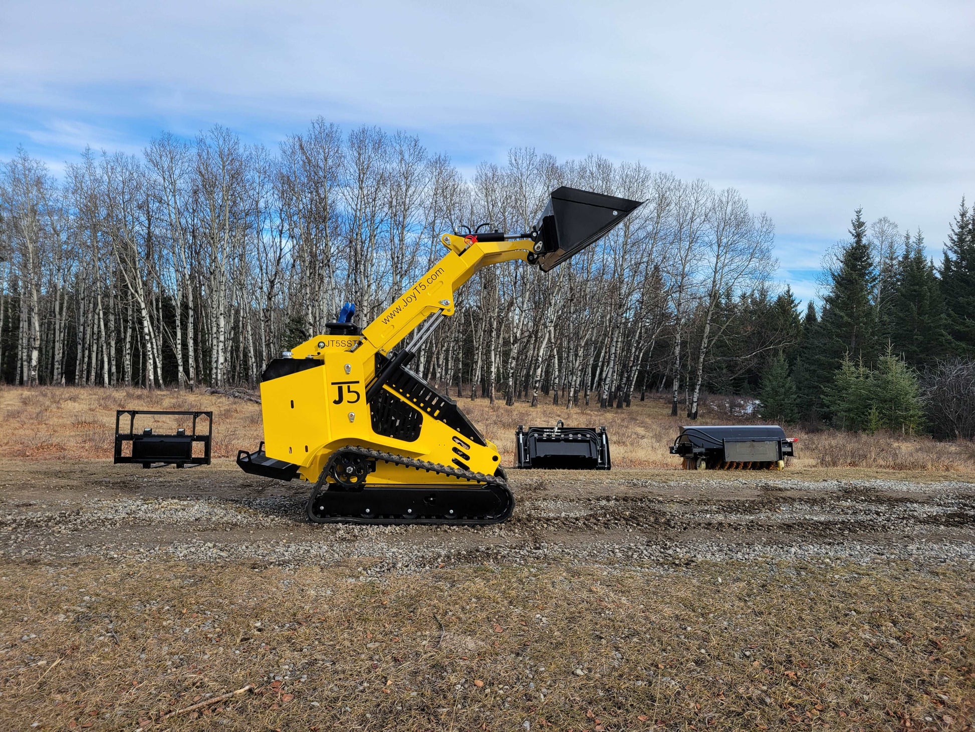JoyT5 tracked mini skid steer loader with raised bucket outdoors, with pallet fork, general-purpose bucket, and sweeper attachments in the background.